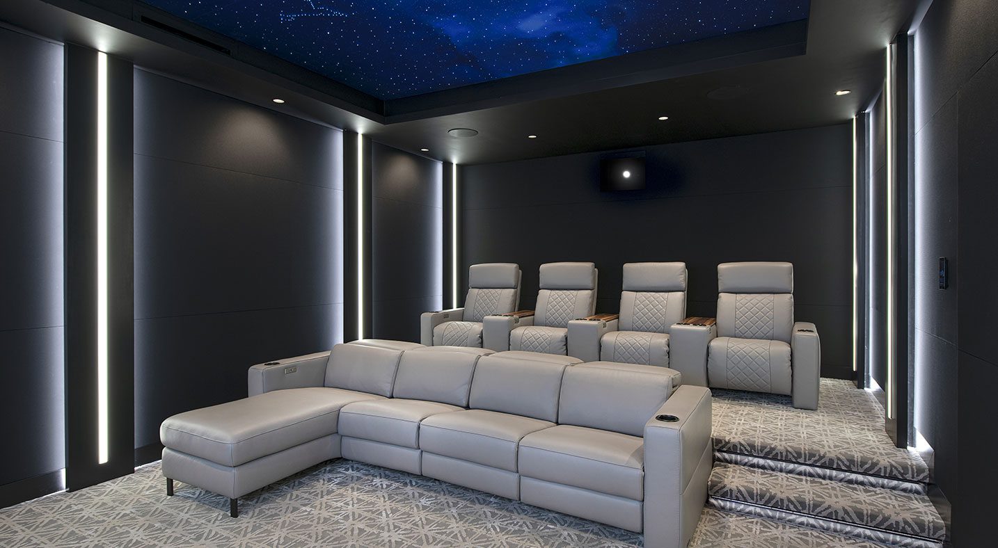 A minimalist home theater with comfortable gray leather recliners in a stadium-style arrangement. The room features a fiber optic star ceiling, creating a celestial atmosphere perfect for a cinematic escape. The vertical light strips on the walls enhance the modern, streamlined design, while the understated color palette contributes to a sleek, refined aesthetic.