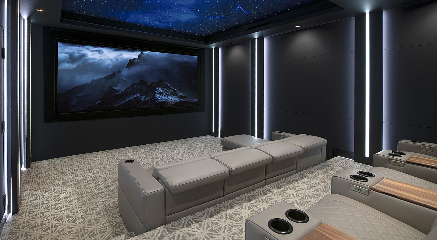 A refined home theater featuring sleek gray leather recliners arranged in rows. The seating includes built-in cup holders with wood accents, offering both functionality and style. The walls are designed with vertical lighting strips that add a modern aesthetic, creating a balanced and well-lit ambiance. A large screen displays a dramatic mountain landscape, complementing the sophisticated decor and enhancing the cinematic experience.