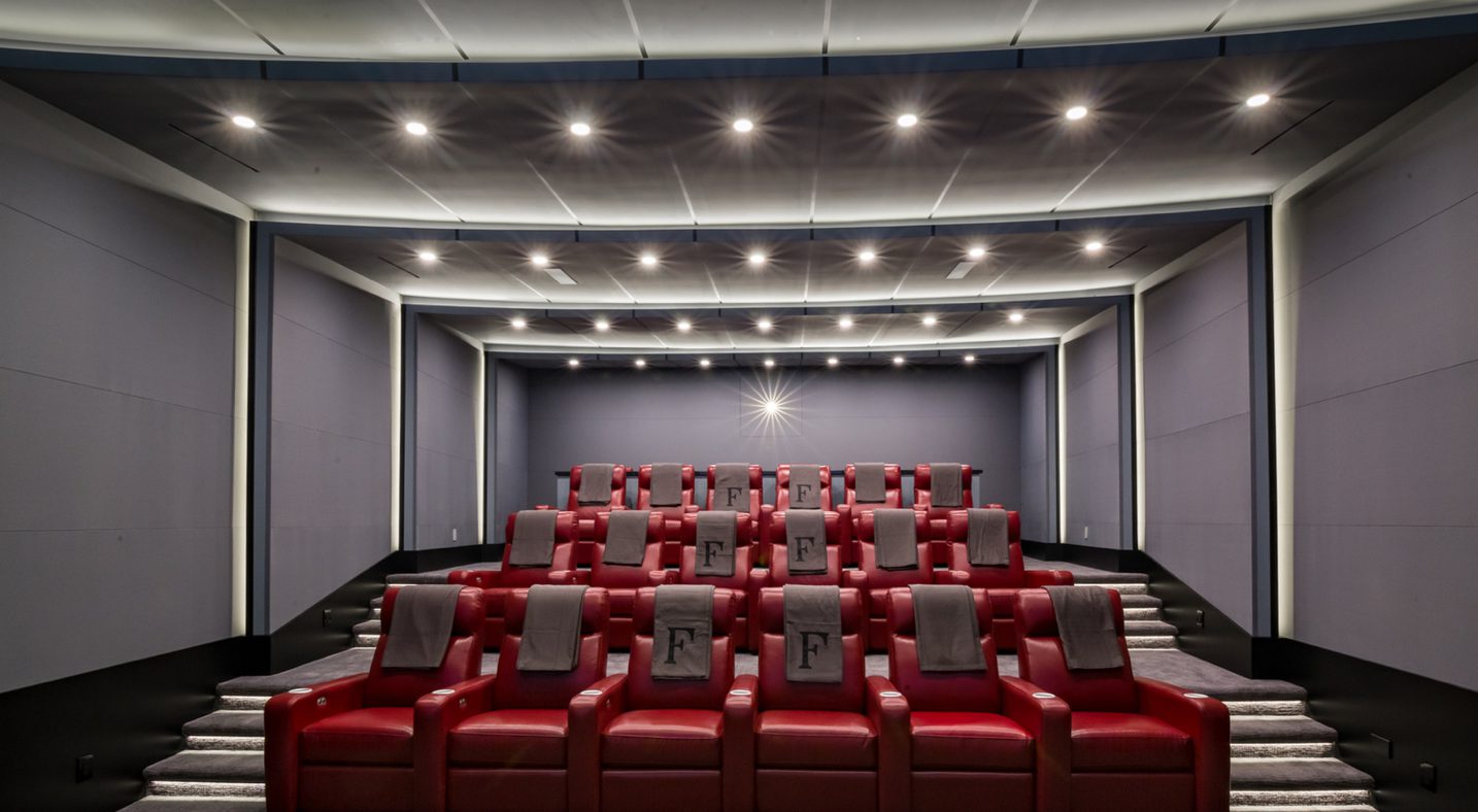 A larger theater space with bold red leather recliners, arranged in multiple rows reminiscent of a traditional cinema. Each seat features custom embroidery on the headrest, adding a personalized touch to the setup. The ceiling lighting is arranged to create a soft glow, illuminating the room without distracting from the screen. This space is designed for larger gatherings, blending luxury and classic theater ambiance.