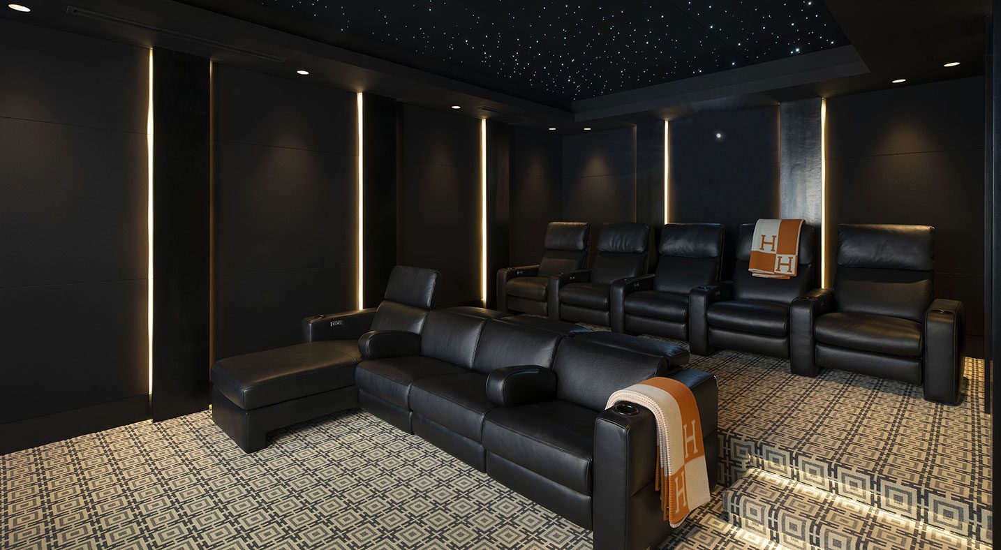 This theater room showcases black leather recliners with plush cushioning, arranged in two elevated rows to optimize viewing angles. The room includes a fiber optic star ceiling, casting a subtle, starlit effect that enhances the immersive experience. The geometric carpet pattern and minimal wall lighting add to the room's luxurious and cozy atmosphere, ideal for high-end home theaters.